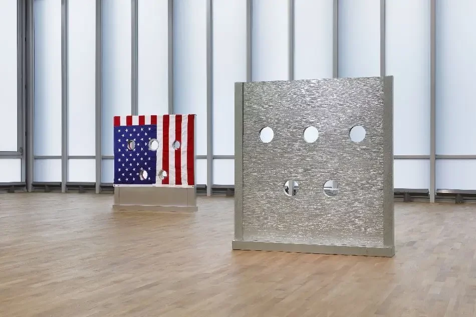 Zwei Pranger, ein silberner im Vordergrund rechts, einer mit der amerikanischen Flagge bedruckter im Hintergrund links in großem Raum