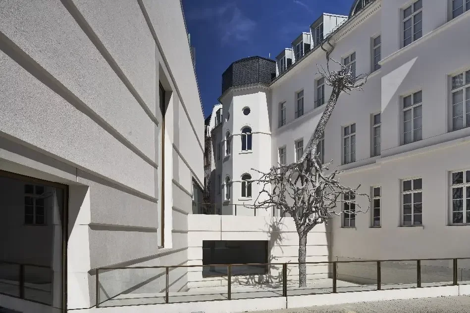 Skulptur mit zwei an den Kronen ineinander verschlungene Bäume, bei denen einer in der Erde verwurzelt ist, während der Stamm und die Wurzeln des anderen in den Himmel ragen auf Platz, Eingang des Neubaus links, rechts Teile des Altbaus