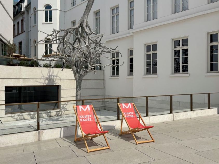 Zwei rote Liegestühle mit der Aufschrift Kunstpause auf einem Platz vor einem Gebäude