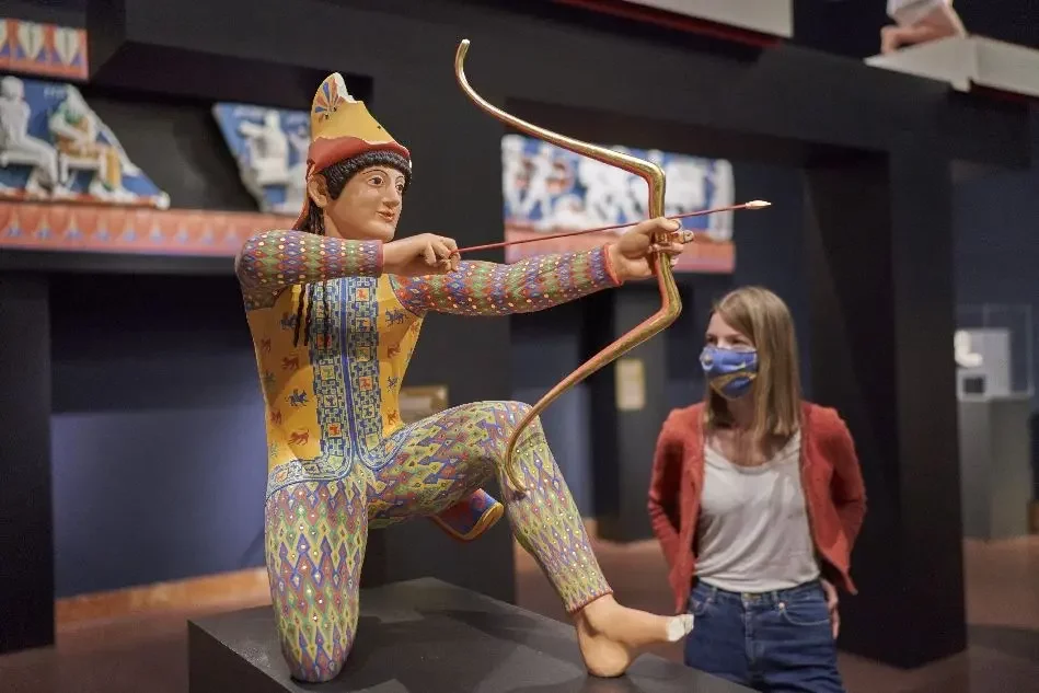 Frau mit Maske betrachtet bunte Skulptur eines knieenden Kriegers mit Pfeil und Bogen in Ausstellung